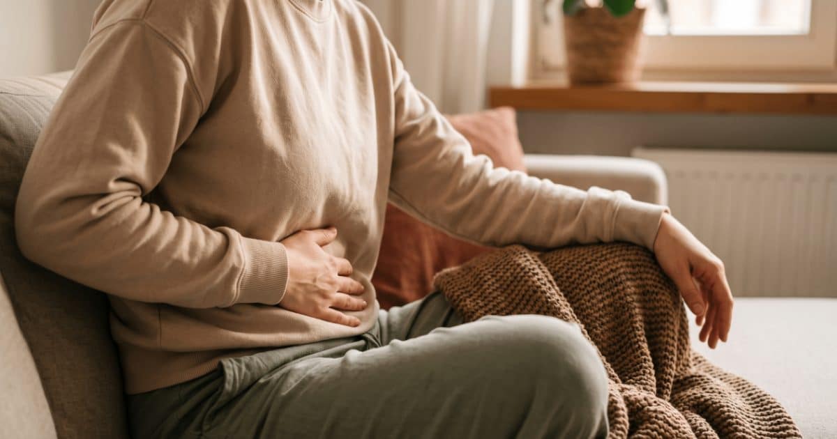 Frau sitzt zusammengezogen auf einem Sofa, eine Hand ruht tief auf dem Unterbauch, eine Strickdecke über den Knien