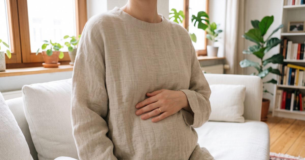 Frau sitzt entspannt auf einem Sofa, eine Hand ruht auf dem Bauch, sie trägt einen weiten Leinenpullover