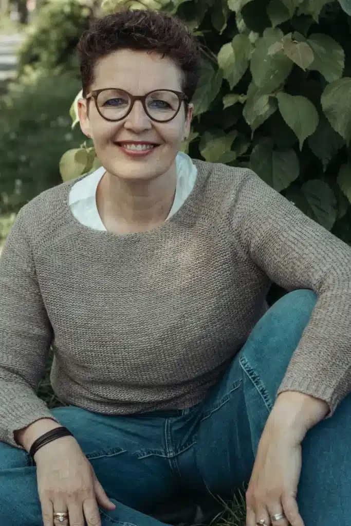 Silke Lemhöfer, Heilpraktikerin und Hormoncoach, sitzt lächelnd vor grünem Blattwerk