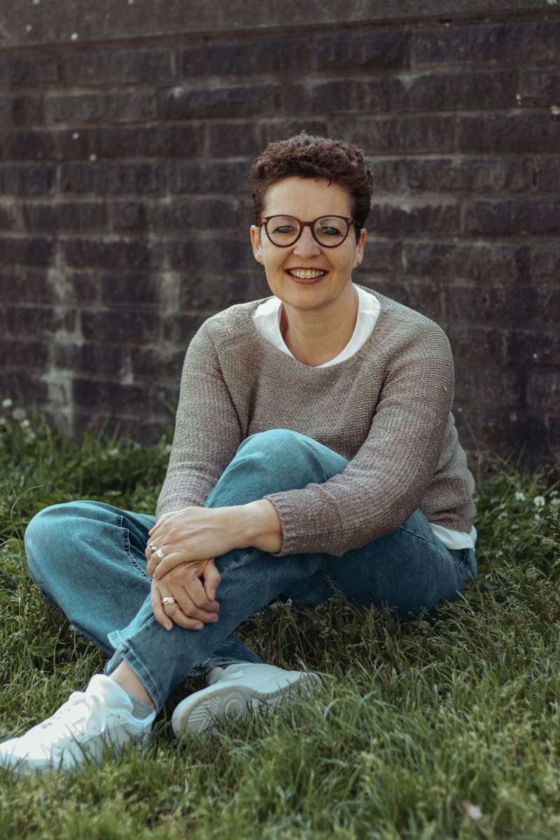 Silke Lemhöfer sitzt im Gras vor einer Backsteinmauer und lächelt